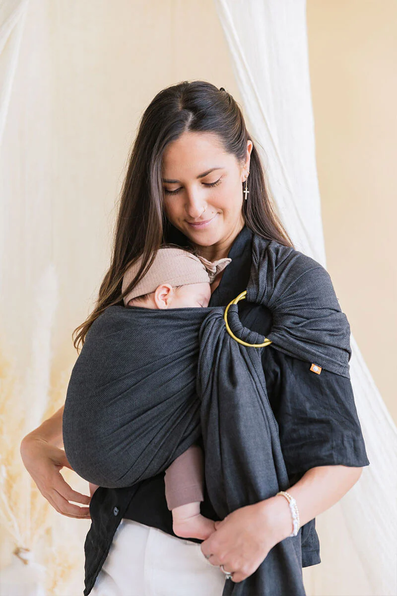Mother wearing her baby in a dark gray Tula Ring Sling with a gold ring, holding the infant close to her chest. The fabric drapes across her shoulder and torso, creating a snug and supportive carry for the newborn.
