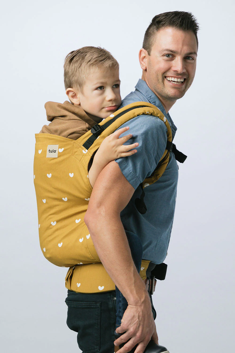 Father carrying his older child in a mustard yellow Tula Preschool Carrier with a white heart print. The wide panel and strong waistband provide support for bigger kids, making it ideal for long walks or outings.
