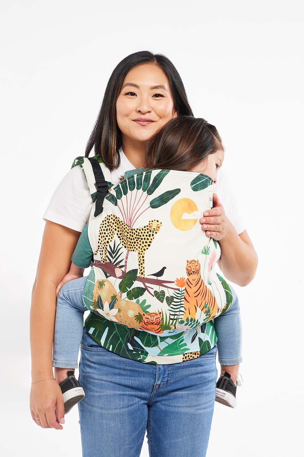Mother carrying her toddler in a colorful Tula Toddler Carrier featuring a jungle print with cheetahs, tigers, and tropical leaves. The structured panel and padded straps keep both parent and child comfortable during extended babywearing.