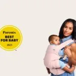 Mother wearing her baby in a light pink Tula Explore Baby Carrier, holding the child close in a front carry. A “Parents Best for Baby 2025” award badge appears beside her, highlighting the carrier’s recognition for comfort and design.