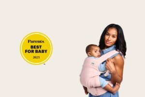Mother wearing her baby in a light pink Tula Explore Baby Carrier, holding the child close in a front carry. A “Parents Best for Baby 2025” award badge appears beside her, highlighting the carrier’s recognition for comfort and design.