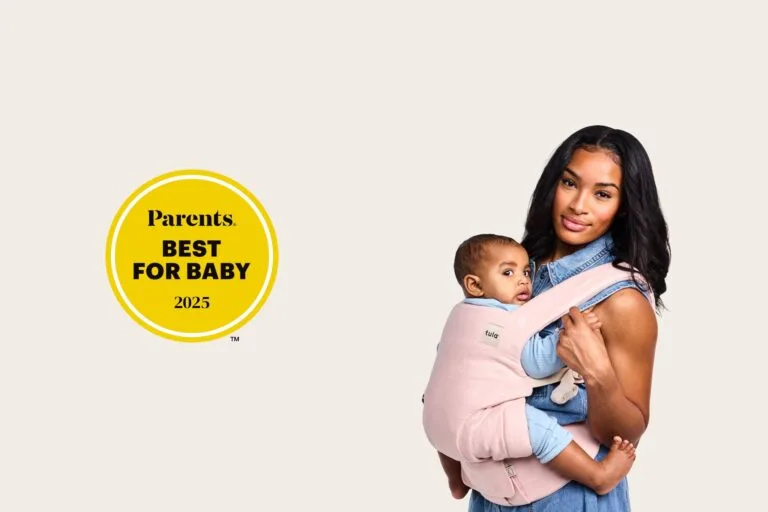 Tula Baby Carrier Review: My Honest Experience and Favorites 2 Mother wearing her baby in a light pink Tula Explore Baby Carrier, holding the child close in a front carry. A “Parents Best for Baby 2025” award badge appears beside her, highlighting the carrier’s recognition for comfort and design.