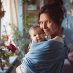 Smiling mom holding her baby in a light blue baby carrier wrap near a sunny window filled with plants and flowers. The image captures cozy, hands-free babywearing wraps ideal for bonding with a newborn at home.