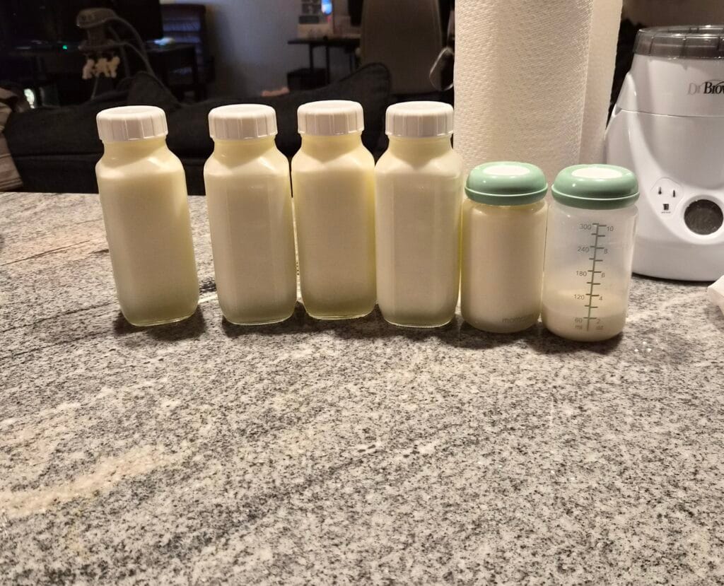 breastmilk-oversupply ix bottles of pumped breast milk lined up on a kitchen counter including four large glass bottles and two Momcozy pump bottles all full