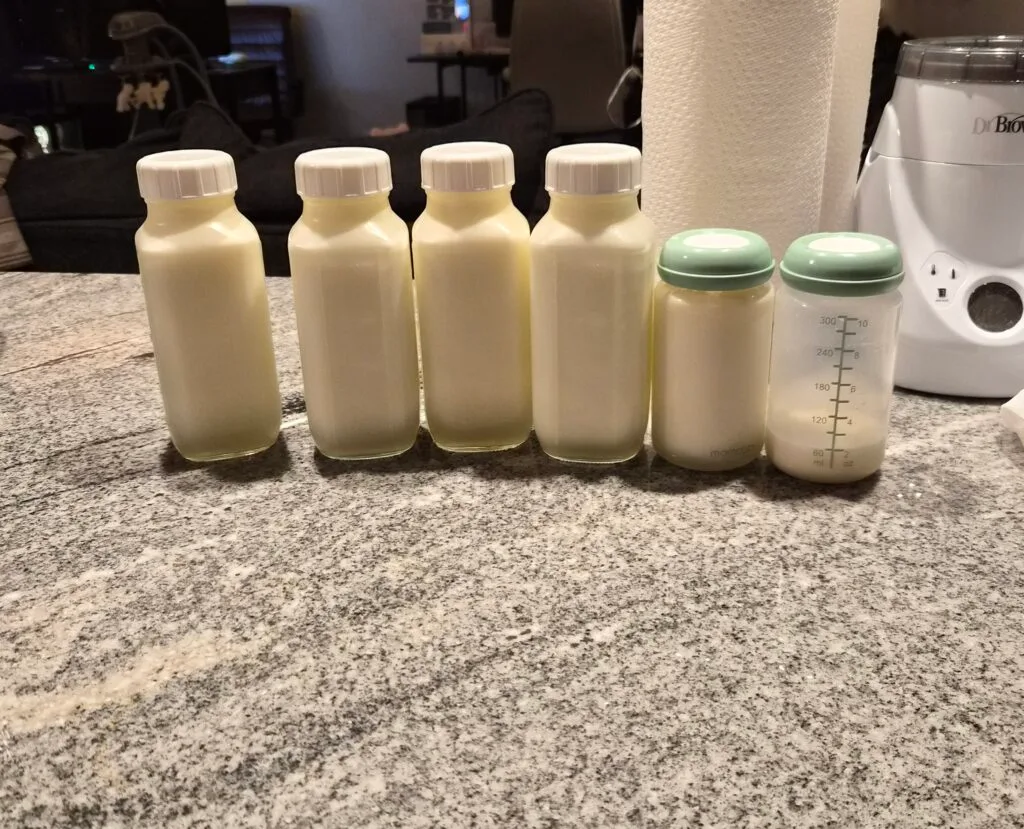 ix bottles of pumped breast milk lined up on a kitchen counter including four large glass bottles and two Momcozy pump bottles all full